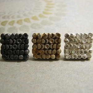 May include: Three square-shaped earrings with a textured surface. The earrings are made of metal and are in black, gold, and silver.