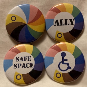Könnte beinhalten: Vier runde Knöpfe mit einem Regenbogenmuster und einer weißen Mitte. Die Knöpfe haben unterschiedliche Texte in der Mitte: "ALLY", "SAFE SPACE", ein Rollstuhlsymbol und ein Kreis mit einem Strich durch.