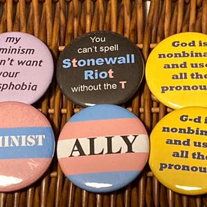 Op de afbeelding: Een verzameling van zes ronde buttons met verschillende teksten. De buttons bevatten zinnen als "my feminism doesn't want your transphobia", "Stonewall Riot" en "God is nonbinary and uses all the pronouns". De buttons zijn in roze, blauw, zwart en geel.