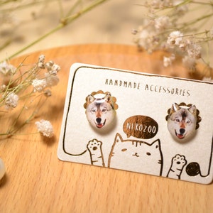 Puede incluir: Un par de pendientes de lobo hechos a mano en una tarjeta blanca con el texto "Handmade Accessories" y "NIKOZO0" en letras doradas. Los pendientes son redondos y presentan la cara de un lobo.