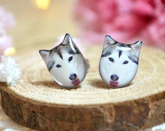 May include: A pair of silver stud earrings featuring a close-up image of a husky dog's face. The earrings are displayed on a wooden surface with pink flowers in the background.