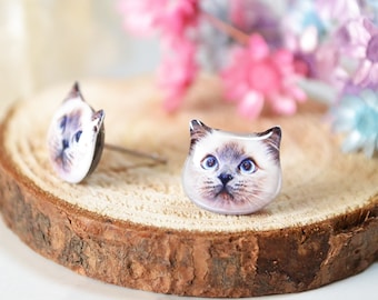May include: A pair of cat-shaped earrings with a white and brown Siamese cat design. The earrings are displayed on a wooden surface with pink and blue flowers.