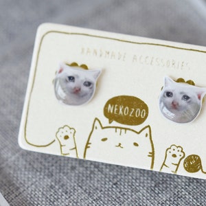 Puede incluir: Un par de pendientes con forma de cara de gato blanco en una tarjeta con el texto "Handmade Accessories" y "NekoZoo".