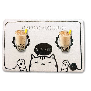 Op de afbeelding: Een witte kaart met zwarte en gele illustraties van een kat, een spreekballon met de tekst "NEKOZOO" en twee glazen bubble tea met rietjes. De kaart heeft bovenaan de tekst "HANDMADE ACCESSORIES".