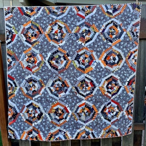 May include: A Halloween-themed quilt with a repeating geometric pattern. The quilt features orange, black, gray, and white fabrics arranged in a spiderweb design. The background is a gray fabric with a small white star pattern. The quilt is hanging on a wooden fence.