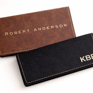 K&ouml;nnte beinhalten: Zwei Visitenkartenetuis aus Leder. Eines ist braun mit dem goldfarbenen Schriftzug "ROBERT ANDERSON". Das andere ist schwarz mit dem goldfarbenen Schriftzug "KBE". Beide haben wei&szlig;e N&auml;hte an den R&auml;ndern.