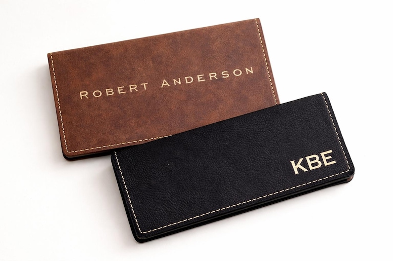 K&ouml;nnte beinhalten: Zwei Visitenkartenetuis aus Leder. Eines ist braun mit dem goldfarbenen Schriftzug "ROBERT ANDERSON". Das andere ist schwarz mit dem goldfarbenen Schriftzug "KBE". Beide haben wei&szlig;e N&auml;hte an den R&auml;ndern.