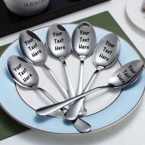 May include: A set of six polished silver-toned metal spoons arranged in a fan pattern on a plate. Each spoon bowl features the black printed text "Your Text Here" centered on the surface, showcasing a smooth, reflective finish and curved handles.