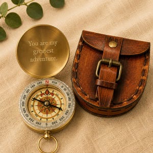 Peut inclure: Une boussole en laiton avec un couvercle à charnière gravé du texte "You are my greatest adventure." La boussole présente un cadran directionnel détaillé avec des points cardinaux et une aiguille. À côté se trouve une pochette en cuir marron.