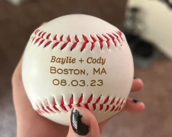 May include: White baseball with red stitching and the text "Baylie + Cody Boston, MA 08.03.23" in gold lettering.