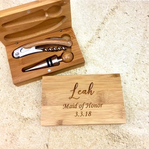 May include: A wooden box set containing a corkscrew and bottle stopper. The box is engraved with the name "Leah", the title "Maid of Honor", and the date "3.3.18".