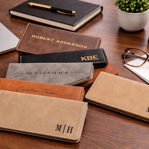 May include: A collection of personalized leather wallets in various colors, including brown, black, and tan, with names or initials embossed on them. The wallets are arranged on a wooden surface, alongside a laptop, notebook, and glasses.
