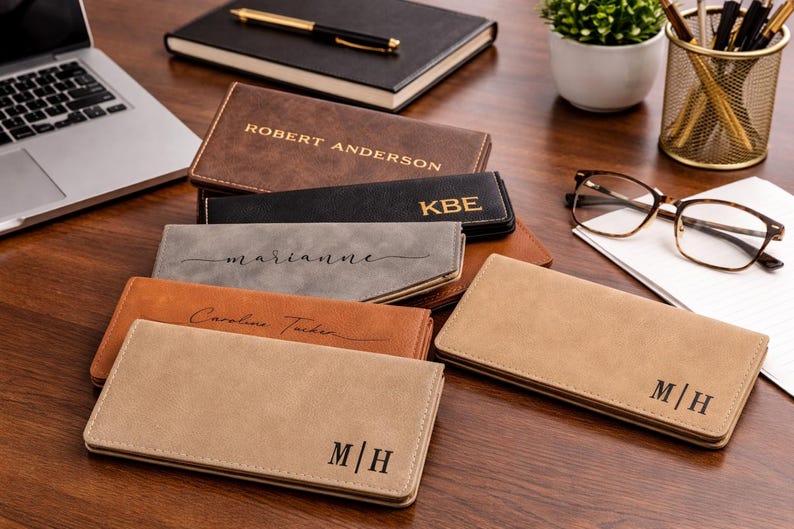 K&ouml;nnte beinhalten: Eine Sammlung personalisierter Ledergeldb&ouml;rsen in verschiedenen Farben, darunter Braun, Schwarz und Beige, mit eingepr&auml;gten Namen oder Initialen. Die Geldb&ouml;rsen liegen auf einer Holzoberfl&auml;che, zusammen mit einem Laptop, Notizbuch und Brille.