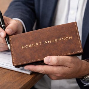 K&ouml;nnte beinhalten: Ein brauner Leder-Scheckbuchumschlag mit dem Namen "ROBERT ANDERSON" in goldfarbenen Buchstaben. Ein schwarzer Stift wird &uuml;ber einem Scheck auf einer Holzoberfl&auml;che gehalten. Der Scheckbuchumschlag ist rechteckig und scheint neu zu sein.