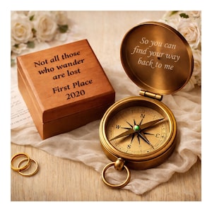 May include: A vintage-style brass compass with an open lid, displaying the text "So you can find your way back to me." A wooden box with the inscription "Not all those who wander are lost, First Place 2020" is next to the compass. Gold rings are also visible.