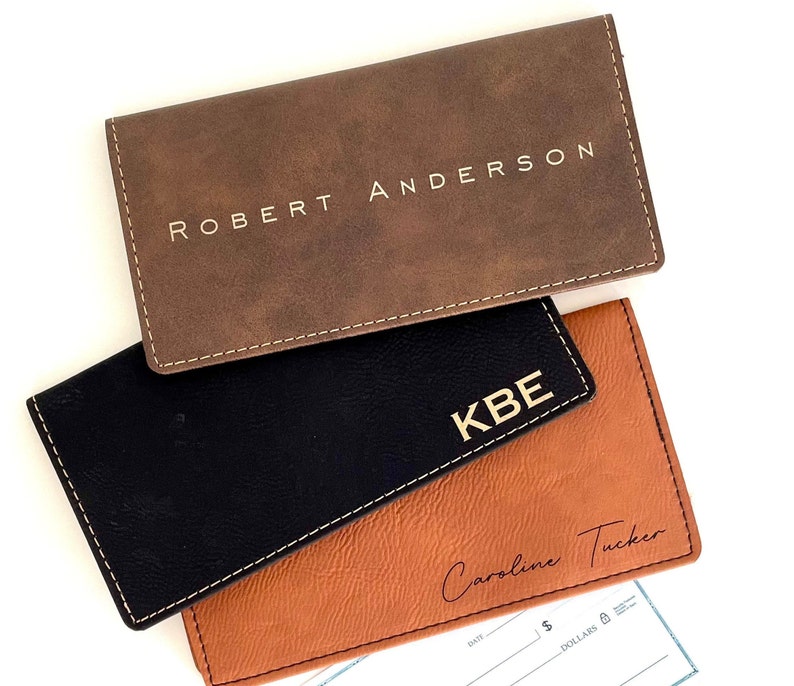 K&ouml;nnte beinhalten: Drei personalisierte Scheckhefth&uuml;llen aus braunem, schwarzem und hellbraunem Leder. Die braune H&uuml;lle ist mit "ROBERT ANDERSON" gepr&auml;gt, die schwarze mit "KBE" und die hellbraune mit "Caroline Tucker". Alle H&uuml;llen haben wei&szlig;e N&auml;hte.