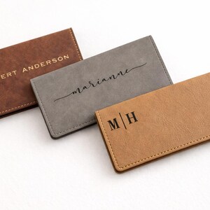 K&ouml;nnte beinhalten: Drei personalisierte Leder-Kartenetuis in Braun, Grau und Beige. Das braune Kartenetui tr&auml;gt den Schriftzug "ROBERT ANDERSON" in goldfarbenen Buchstaben. Das graue Kartenetui tr&auml;gt "Marianne" in Schreibschrift. Das beige Kartenetui tr&auml;gt "M | H".