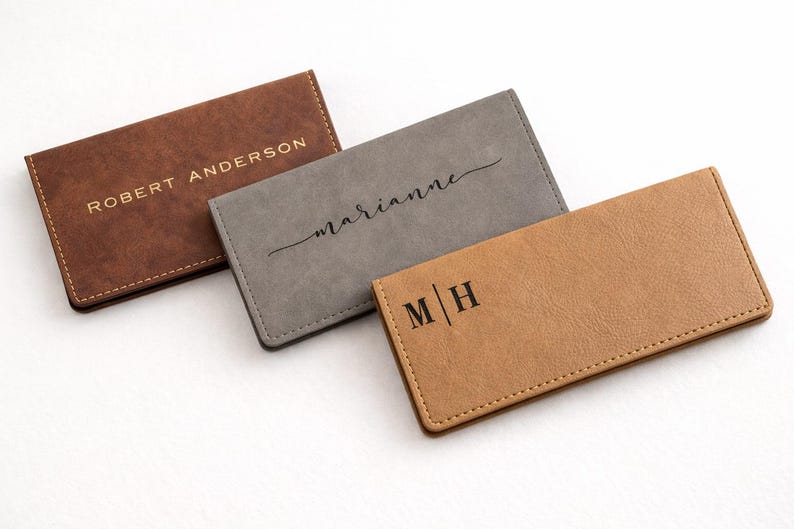 K&ouml;nnte beinhalten: Drei personalisierte Leder-Kartenetuis in Braun, Grau und Beige. Das braune Kartenetui tr&auml;gt den Schriftzug "ROBERT ANDERSON" in goldfarbenen Buchstaben. Das graue Kartenetui tr&auml;gt "Marianne" in Schreibschrift. Das beige Kartenetui tr&auml;gt "M | H".
