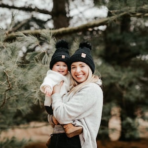 Puede incluir: Una mujer y un niño llevan gorros de punto negros a juego con pompones. La mujer sostiene al niño en sus brazos. Ambos sonríen y miran a la cámara. El fondo es un bosque verde borroso.