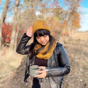 May include: A person wearing a mustard yellow knit beanie, a mustard yellow infinity scarf, a black leather jacket, and black jeans is holding a gray ceramic mug. The person is standing in a wooded area with fall foliage in the background.