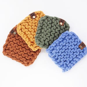 Könnte beinhalten: Vier handgestrickte Becherhüllen in Braun-, Gelb-, Grün- und Blautönen. Jede Hülle hat ein strukturiertes, gewebtes Design und ein kleines braunes Lederetikett mit einem Knopf. Die Hüllen sind auf weißem Hintergrund angeordnet.