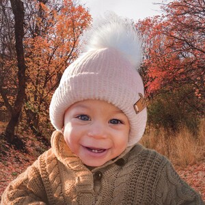 May include: A baby wearing a pink knit beanie with a white pom pom and a brown cable knit sweater. The baby is smiling and looking at the camera. The background is a blurry image of fall foliage.
