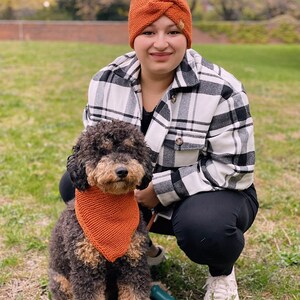 May include: A woman wearing a black and white plaid shirt and an orange headband is squatting down with a brown and black dog wearing an orange bandana. The dog is on a leash.