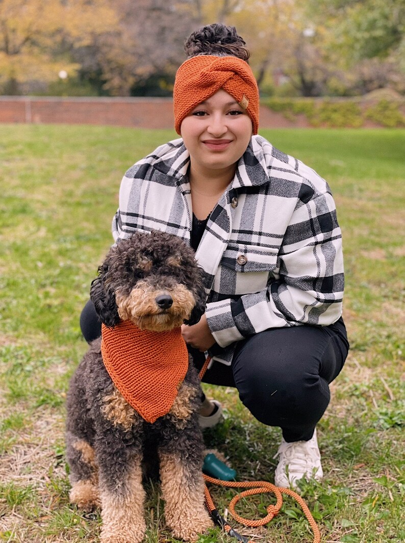 May include: A woman wearing a black and white plaid shirt and an orange headband is squatting down with a brown and black dog wearing an orange bandana. The dog is on a leash.