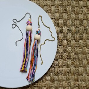 May include: Two colorful tassel ceiling fan pulls. Each tassel features a wooden bead, a patterned bead, and a bundle of yarn in shades of purple, pink, yellow, orange, and blue. The pulls have metal chains.