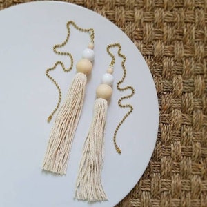 May include: Two white and beige tassel pull chains with gold beads. The tassels are made of white cotton yarn and the beads are round and wooden.