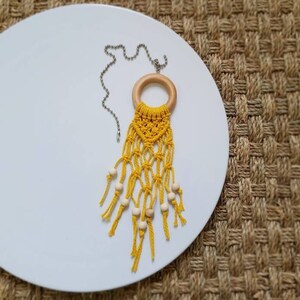 May include: A yellow macrame hanging with wooden beads and a wooden ring, attached to a silver chain. The hanging is on a white plate.