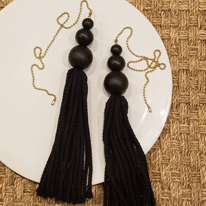 May include: Two black tassel pulls with gold chains and black wooden beads. The tassels are made of black yarn and are hanging from a white plate.