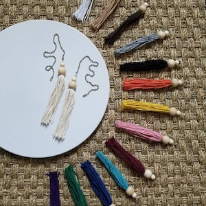 May include: A collection of 14 colorful tassel pulls with wooden beads. The tassels are made of various colors, including white, beige, brown, gray, black, orange, yellow, pink, maroon, turquoise, blue, green, and purple. The tassels are arranged in a semi-circle on a woven surface.