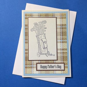 May include: A greeting card featuring a cream-colored center panel with a black ink illustration of a golf bag filled with clubs and three golf balls. The card has a tan and blue plaid background, a light blue border, and the text Happy Father's Day.
