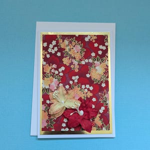 May include: A handmade greeting card with a red floral pattern and a gold border. Two paper flowers, one yellow and one red, are attached to the front. The card is set against a light blue background.