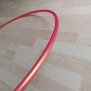 Puede incluir: Un aro de hula rojo vibrante, parcialmente visible, sobre un suelo de madera clara. La forma curva del aro sugiere un artículo de fitness o recreativo. El color es un rojo brillante y sólido.