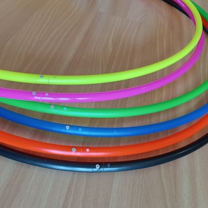 Puede incluir: Seis aros de hula de plástico de colores apilados uno encima del otro. Los aros son negros, naranjas, azules, verdes, rosas y amarillos.