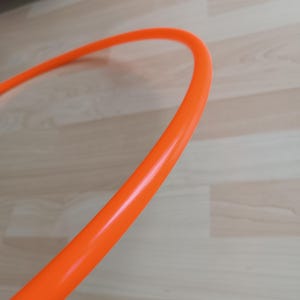 Puede incluir: Hula hoop de color naranja brillante. La sección curva del aro está en primer plano, con un suelo de madera en el fondo. El aro es de un color vibrante y sólido, y parece estar hecho de plástico.