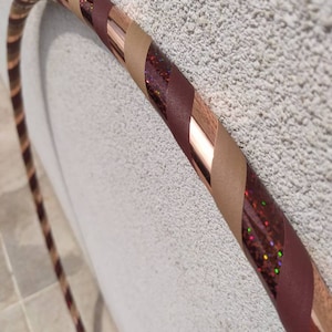 Puede incluir: Un aro con un diseño a rayas de cobre, marrón y beige. El aro está decorado con purpurina iridiscente.