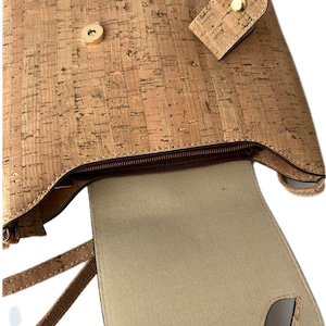 May include: A cork handbag with a brown zipper and a brown strap. The bag is open and the inside is lined with a beige fabric.