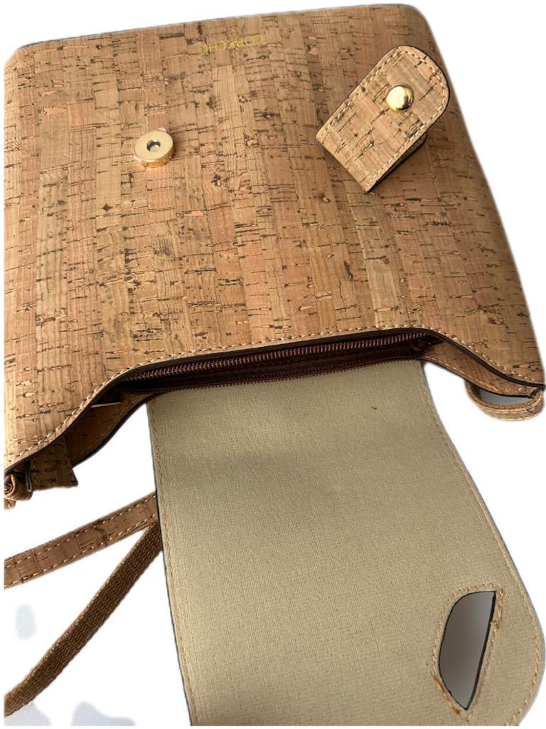 May include: A cork handbag with a brown zipper and a brown strap. The bag is open and the inside is lined with a beige fabric.