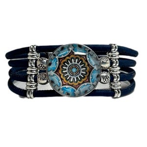 May include: A multi-strand bracelet featuring four dark blue leather cords held together by silver-toned metal spacers. A central circular glass cabochon displays a detailed mandala-like pattern in shades of blue, orange, and white.