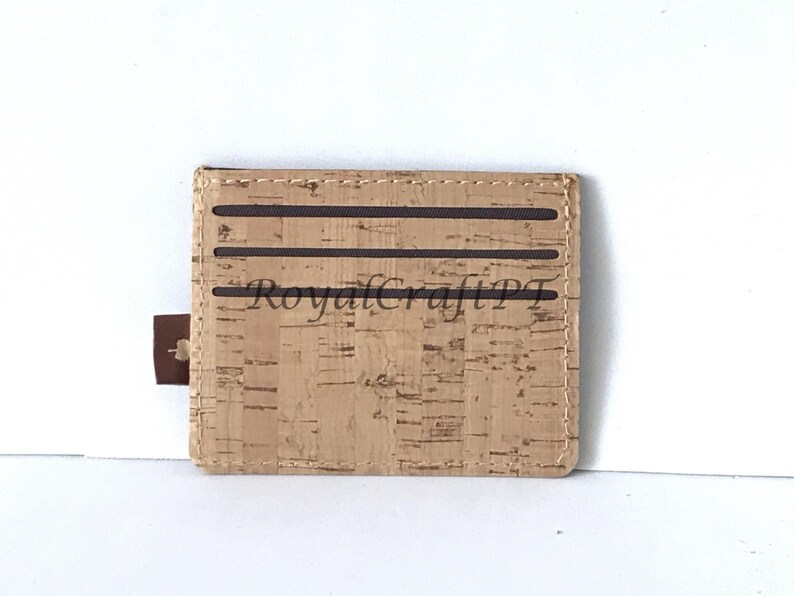May include: A brown cork wallet with a brown stitched border and three brown elastic bands. The wallet has the text "RoyalCraftPT" printed on the front.