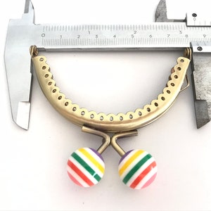 May include: A gold-toned metal purse frame with a scalloped edge, measuring approximately 4 inches in diameter. The frame has two colorful striped balls attached to the bottom.