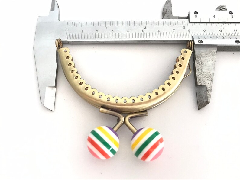 May include: A gold-toned metal purse frame with a scalloped edge, measuring approximately 4 inches in diameter. The frame has two colorful striped balls attached to the bottom.