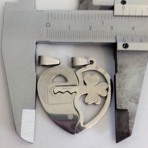 May include: Two silver heart-shaped pendants, one with a keyhole and the other with a four-leaf clover, are measured with a caliper. The pendants are designed to fit together.