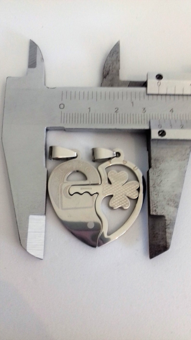 May include: Two silver heart-shaped pendants, one with a keyhole and the other with a four-leaf clover, are measured with a caliper. The pendants are designed to fit together.