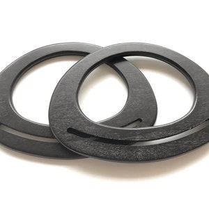 Könnte beinhalten: Zwei schwarze Holzringe, jeder mit einer kreisförmigen Öffnung, sind ineinander verschlungen. Die Ringe haben eine glatte, leicht strukturierte Oberfläche.