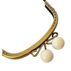 May include: An antique brass-coloured purse frame with small, evenly spaced holes along the edges. Two off-white, spherical beads are attached to the frame with decorative metalwork. The frame is set against a white background.