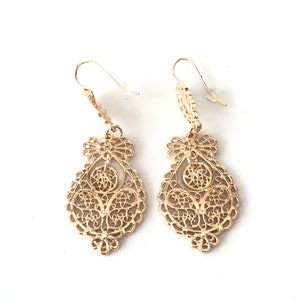 May include: A pair of gold filigree dangle earrings with a delicate, intricate design. The earrings are teardrop shaped and feature a small, round accent at the top.